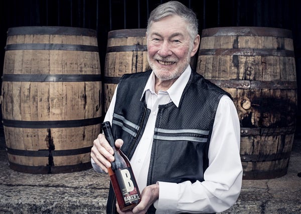 Baker Beam holding a bottle of Baker's in front of 3 bourbon barrels